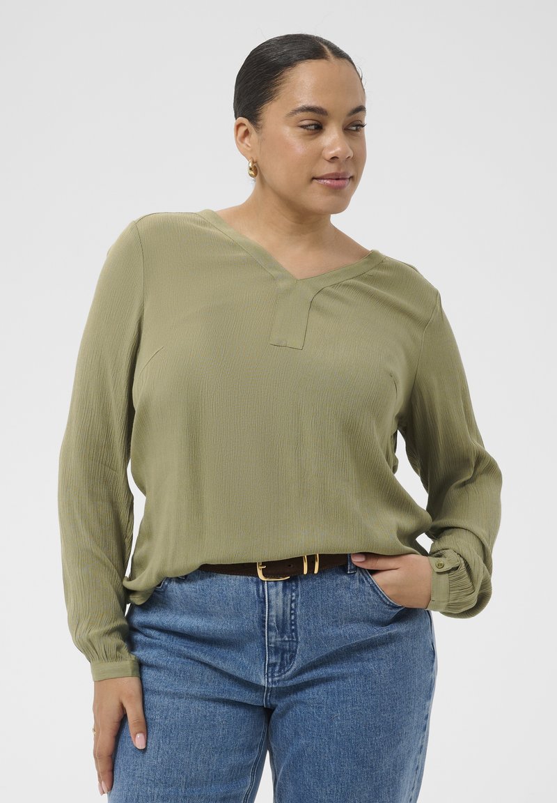 Femme aux cheveux foncés portant un chemisier à manches longues vert olive et un jean bleu, posant avec une main dans la poche et l'autre détendue sur le côté.