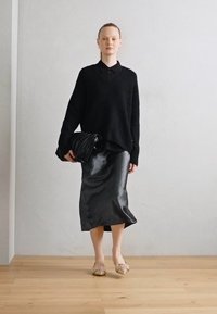 Un pull noir surdimensionné, une jupe midi noire brillante et des chaussures texturées de couleur claire. Le modèle tient un sac plissé noir. Arrière-plan neutre.