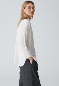 Blusa bianca a maniche lunghe in un tessuto leggero, con una vestibilità comoda e polsini larghi, abbinata a pantaloni scuri e testurizzati. Sfondo neutro.
