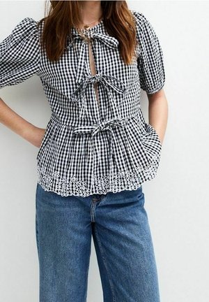 Mujer vistiendo una blusa de cuadros blancos y negros con mangas abullonadas, tres lazos en la parte delantera, dobladillo con ojales y jeans azules de talle alto.