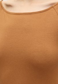 Gros plan d'une personne portant un haut marron uni avec un col rond, montrant le haut de la poitrine et la zone du cou.