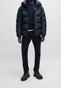 Homme portant une doudoune bleu marine sur un pull foncé et un jean, associée à des bottes noires à lacets, debout devant un fond uni.