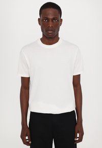 T-shirt blanc à manches courtes, col rond, en coton doux, avec un logo gris clair sur la poitrine, associé à un pantalon noir.