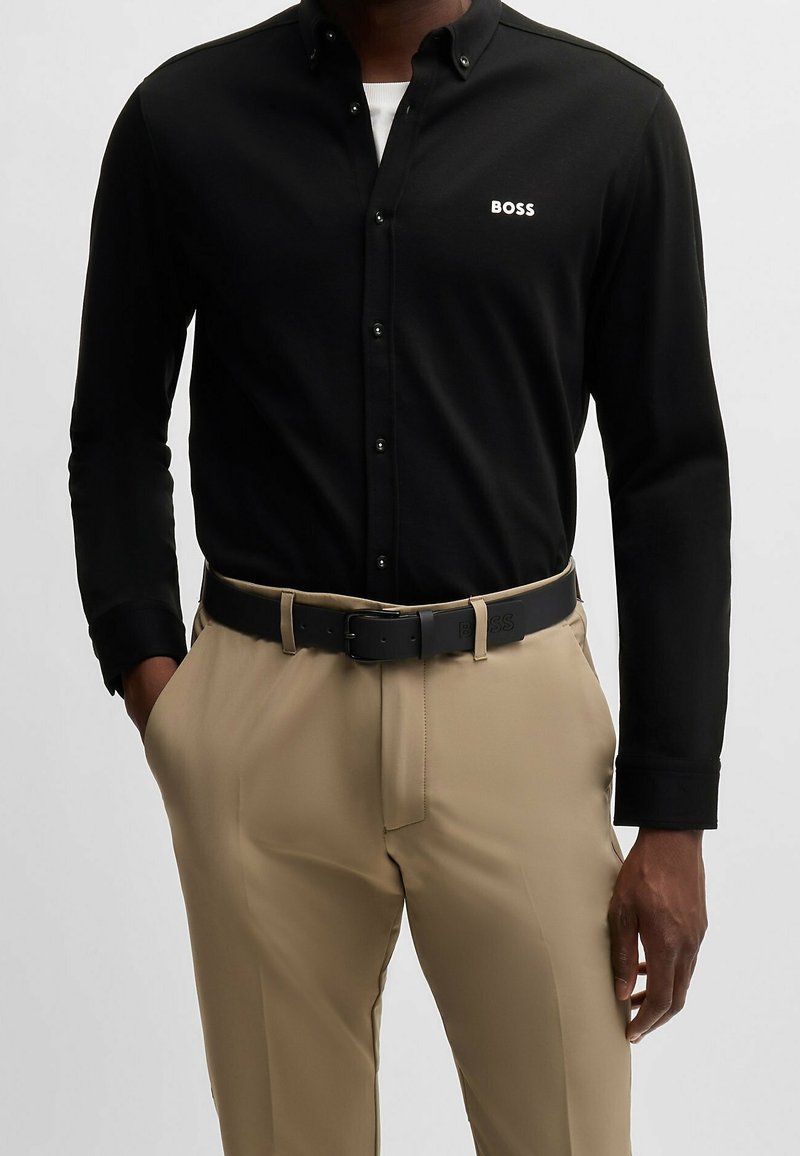 Homme portant une chemise noire à boutons avec le logo "BOSS" sur la poitrine, un pantalon beige et une ceinture noire avec le même logo "BOSS". Mains dans les poches.