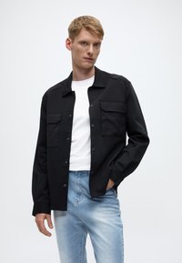 Svart skjortjacka med två bröstfickor, kombinerad med en vit t-shirt och ljusblå avsmalnande jeans, mot en neutral bakgrund.