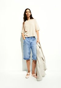Beige gerippter Strickpullover, blaue Jeansshorts und hellrosa Schuhe. Hält ein hellgraues Blazer. Schlichtes Design mit geraden Linien und lässiger Passform.