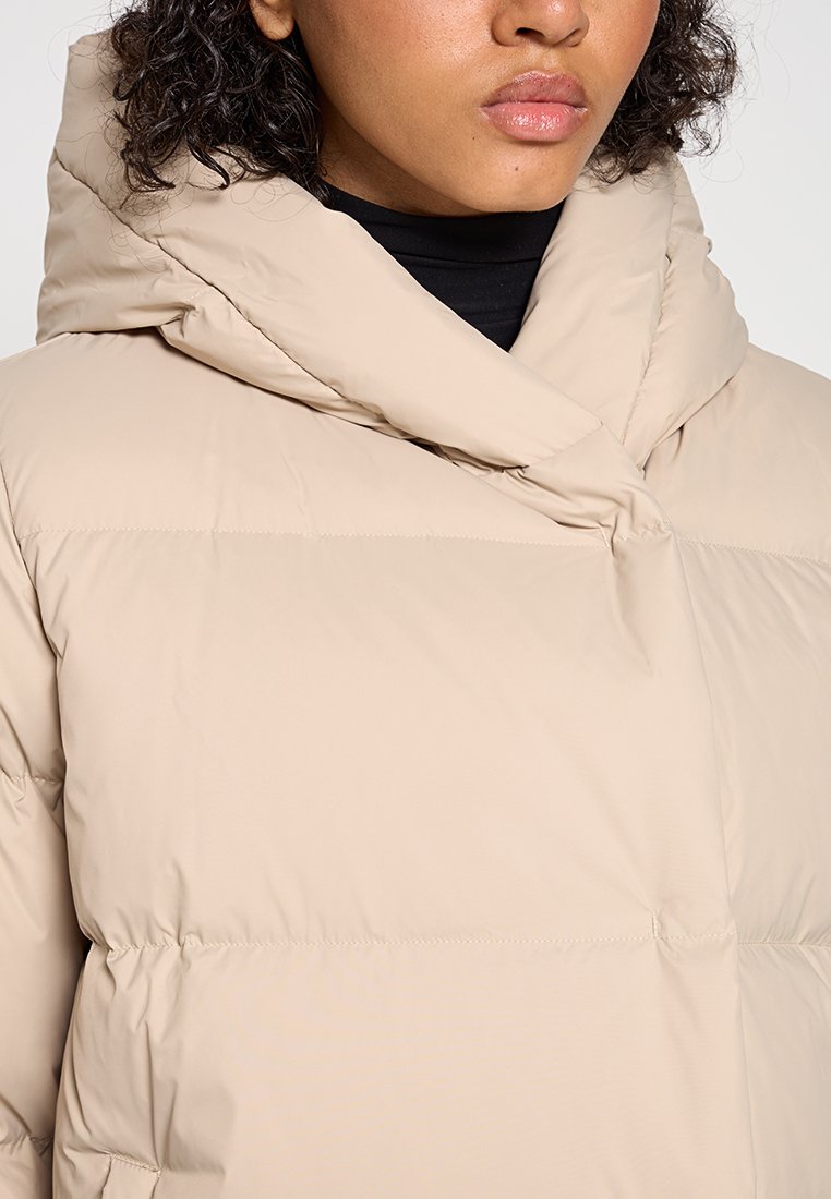 Beżowa kurtka puchowa z wysokim kołnierzem i miękkim, teksturowanym materiałem. Posiada poziome przeszycia oraz luźny krój dla zapewnienia ciepła i komfortu.