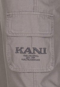 Šedá textilní kapsa s vyšívaným logem "KANI", má detaily stehu a klopové uzavření. Materiál vypadá lehce a odolně.
