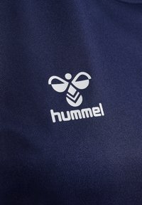 Hvit Hummel-logo med stilisert bie og tekst på mørk blått tekstilbakgrunn.