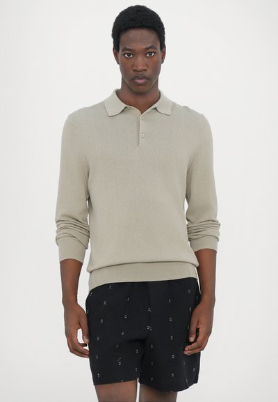 Beige Langarm-Polohemd mit Kragen, gerippten Bündchen und figurbetonter Passform, kombiniert mit schwarzen Shorts mit einem subtilen Muster.