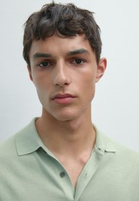 Polo shirt verde chiaro con dettagli dei bottoni, tratti del viso del modello in primo piano, capelli corti scuri, sfondo neutro.