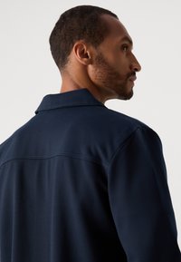 Side profile of a man with short beard wearing a dark navy jacket, looking to the right against a plain light background.