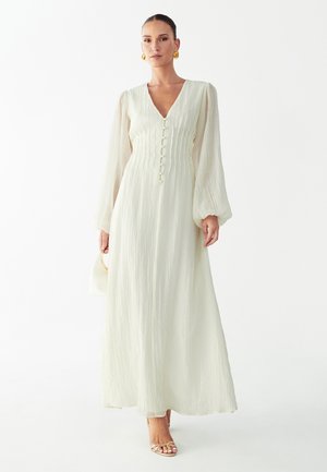 Woman wearing a long, cream, textured dress with long puff sleeves, front buttons, holding a small white handbag, and gold hoop earrings.