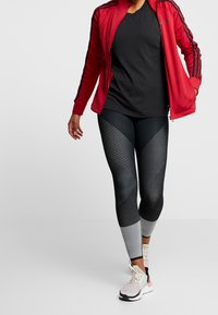 Chaqueta atlética roja con franjas negras, camiseta negra, leggings negros y grises con patrón, y zapatos deportivos blancos con acentos en tonos pastel.