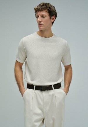 Homme aux cheveux bouclés portant une chemise à manches courtes beige clair et un pantalon blanc cassé, les mains dans les poches sur un fond gris uni.