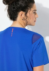 Žena s kudrnatými vlasy staženými do drdolu, která nosí modré sportovní tričko s texturačním vzorem, síťovanými panely na ramenou a malým červeným logem na zádech.