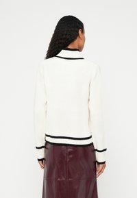 Bela rebrasta jopica s črnimi poudarki na ovratniku in robu, z dolgimi rokavi in standardnim krojem, združena z bordo krilom.