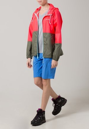 Persona con chaqueta roja y oliva, camiseta gris, pantalones cortos azules y botas de senderismo negras, de pie con una pierna ligeramente adelantada.