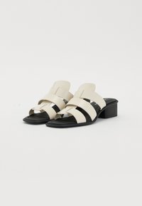 Sandales à talons blocs beiges avec plusieurs brides croisées sur une semelle noire, présentant une finition en cuir lisse et un design à dos ouvert.