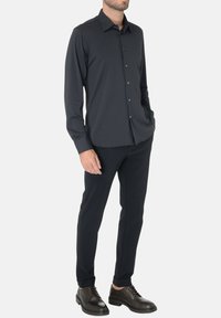 Camicia a maniche lunghe grigia scura con chiusura a bottoni, dotata di colletto classico, indossata con pantaloni scuri e scarpe stringate marroni.