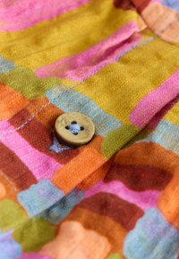 Holzknopf, mit blauem Faden auf buntem Stoff mit abstrakten Mustern in Pink, Orange, Gelb, Grün und Blau genäht.