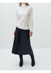 Kobieta w białym swetrze z wysokim kołnierzem, czarnej midi spódnicy i czarnych botkach na obcasie stojąca na tle jasnego, jednolitego tła.