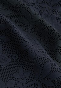 Tissu bleu foncé avec un motif complexe de dentelle florale et de petits trous ouverts, révélant la texture et les détails en relief dans des plis doux.