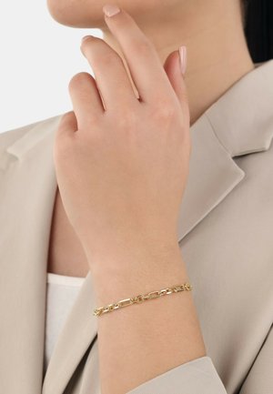Gouden kettingarmband om een pols, met verlengde schakel, een gladde textuur en een veilige sluiting. Draagt een persoon in een blazer in neutrale tinten.
