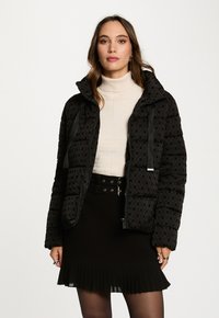 Sort polstret jakke med en quiltet design, der har et gentaget logomønster, lynlås lukning og justerbare snore i hætten.