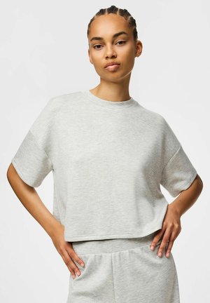 Jonge vrouw met gevlochten haar, gekleed in een lichtgrijze korte mouw crop top en bijpassende broek, staand met handen in de zij.