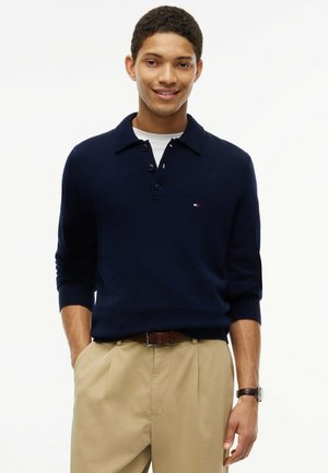 Jonge man die lacht, draagt een marineblauwe trui over een wit overhemd, beige broek en een bruine riem, met één hand in zijn zak.
