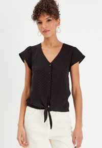 Schwarze Bluse mit kurzen Ärmeln, V-Ausschnitt, Knopfleiste vorne, strukturierte Musterung und einem geknoteten Detail am Saum.
