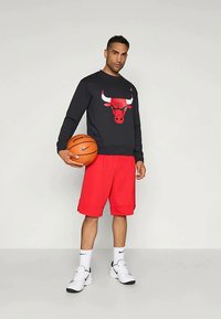 Sweatshirt preta com um logótipo vermelho dos Chicago Bulls, combinada com calções vermelhos. Tênis brancos e segurando uma bola de basquete. Estilo para vestuário atlético.