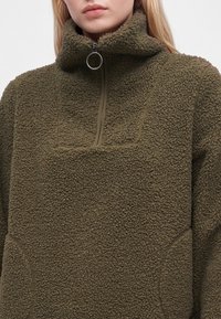 Femme portant un pull en polaire marron avec un col montant et une demi-fermeture éclair avec une grande tirette ronde.