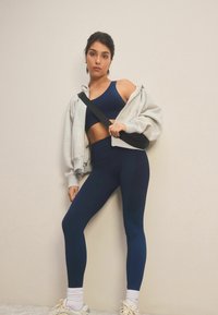 Meresinine spordirinnahoidja, tekstuuri mustriga legginsid, hall tõmblukkidega kapuutsiga jakk, must õlakott, valged sokid ja treeningkingad. Minimalistlik spordivorm.