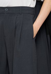 Σκούρο γκρι παντελόνι Tailored με πιέτες μπροστά και πλαϊνές τσέπες, ένα χέρι μέσα σε τσέπη, φορεμένο με λευκό πουκάμισο που έχει μπει μέσα στο παντελόνι.