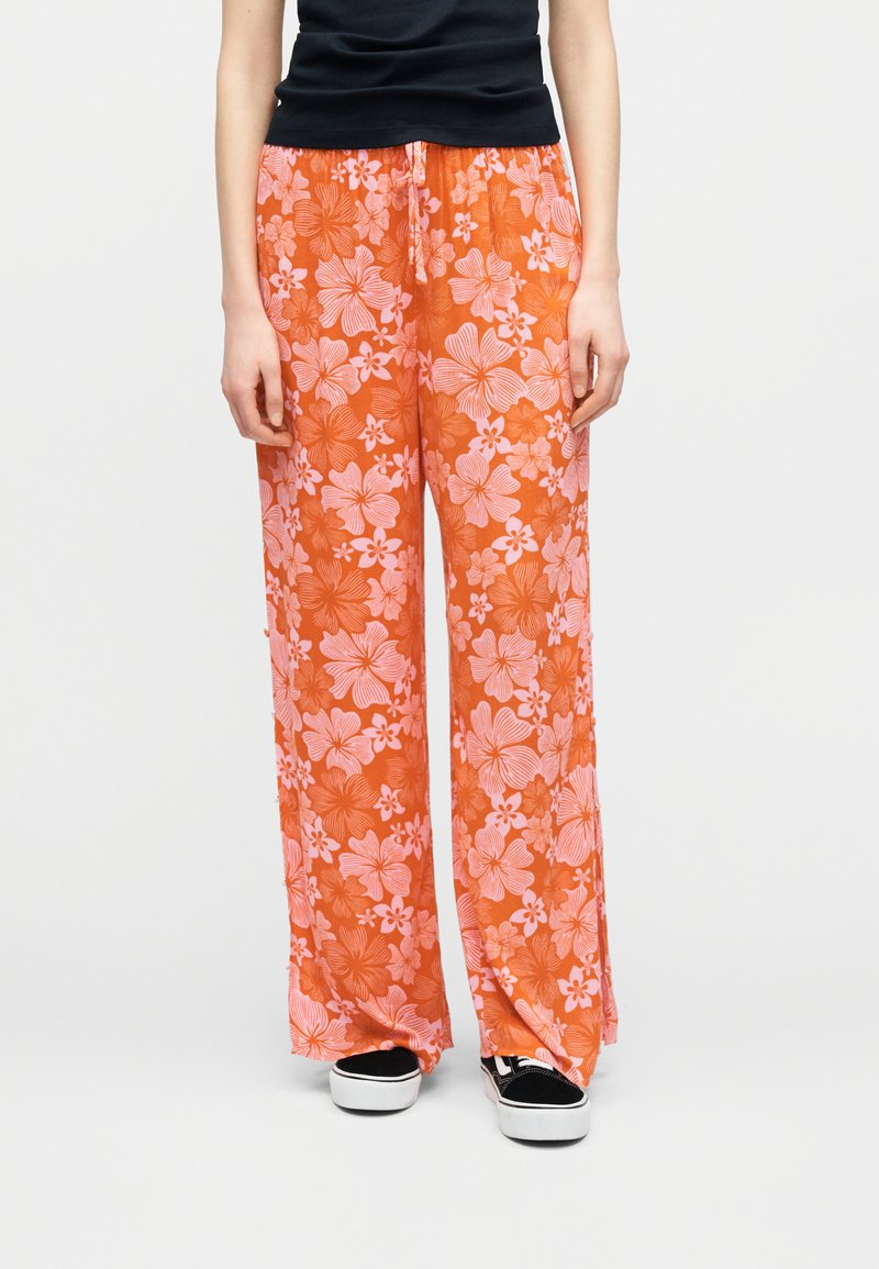 Personne portant un pantalon ample orange à imprimé floral rose, un haut noir et des baskets noir et blanc, debout devant un fond uni.