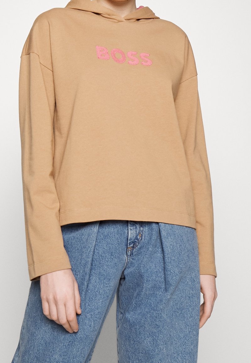 Persona che indossa una felpa con cappuccio beige con scritta rosa "BOSS" e jeans in denim blu a taglio largo, su uno sfondo neutro.