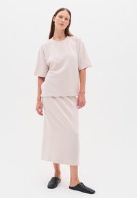 Kvinde iført en løstsiddende lys lyserød kortærmet top og matchende lang nederdel med sorte slip-on sko på hvid baggrund.