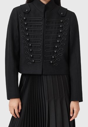 Black military-style cropped jacket with decorative braided loops and buttons, worn over a black pleated skirt.