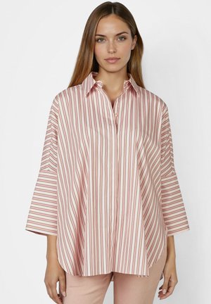 Frau trägt ein lockeres, langärmeliges Hemd mit vertikalen roten und weißen Streifen, kombiniert mit hellrosa Hosen, stehend vor einem schlichten Hintergrund.