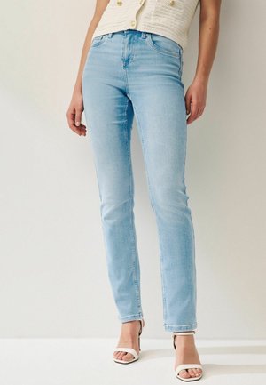 Femme portant un jean slim bleu clair, des sandales blanches à talons ouvertes à l’avant, et un haut sans manches texturé crème avec des boutons.