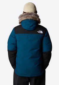Teal- och svart puffjacka med pälsfoder på huvan, med logotyp på ryggen. Tyget ser slätt och isolerat ut, med en avslappnad passform.