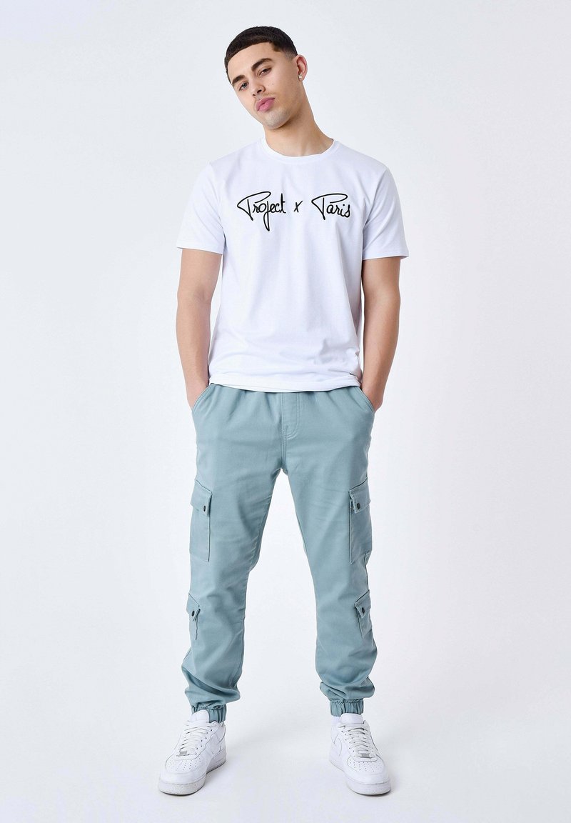 Witte katoenen t-shirt met zwart schrift "Project x Paris" logo, gecombineerd met lichtblauwe cargobroek met zijzakken en elastische boorden. Witte sneakers.