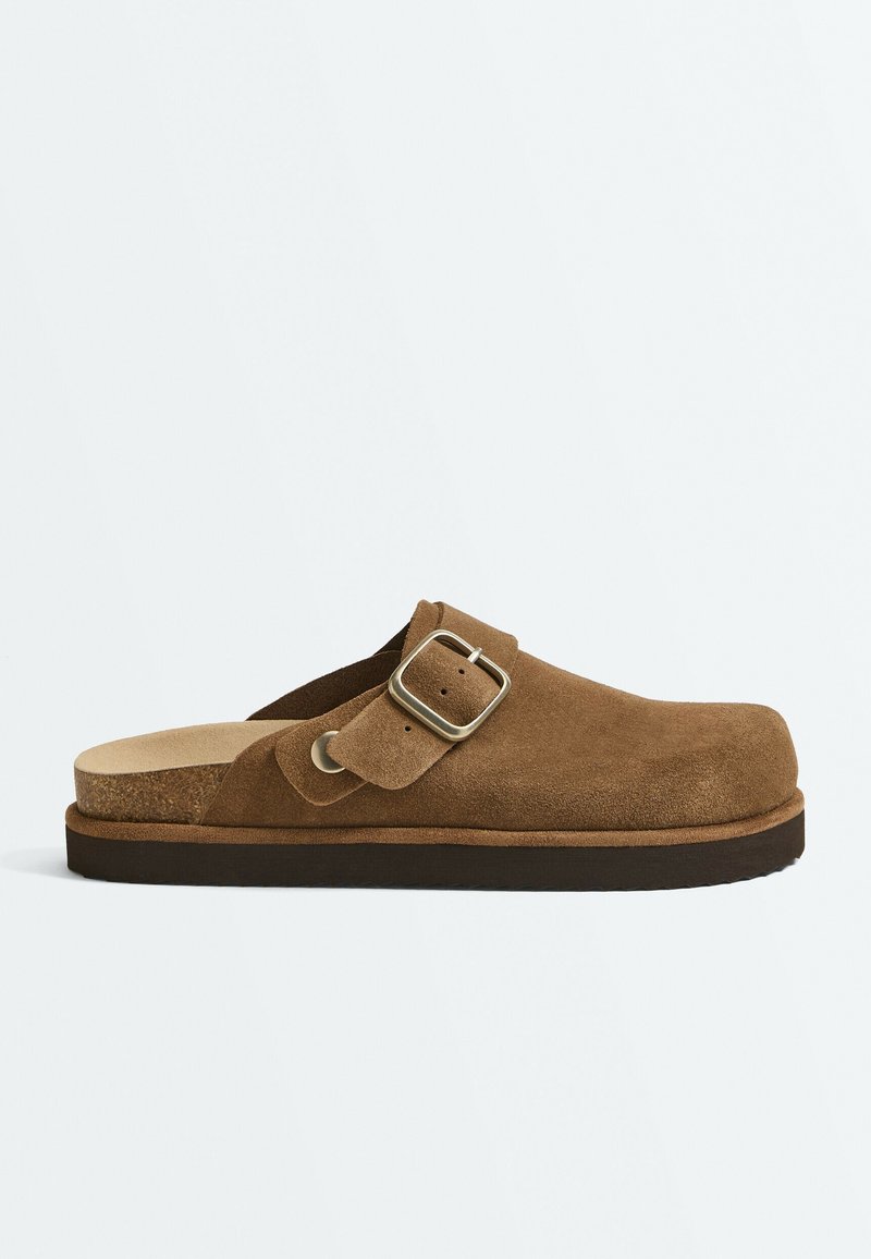 Brown suede slip-on clog with adjustable metal buckle strap and contoured cork sole on a white background.