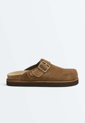 Sabot en daim marron sans lacets avec une bride réglable à boucle métallique et une semelle contournée en liège sur fond blanc.