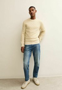 Crème gebreide trui met een textuurpatroon, gecombineerd met lichtblauwe jeans en beige sneakers. Eenvoudig ontwerp met geribbelde manchetten en zoom.