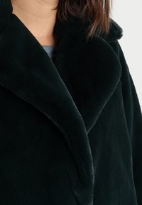 Tummanvihreä tekoturkistakki, jossa on leveä kaulus, pehmeä koostumus ja lovitettu kaulusmalli. Saumojen yksityiskohtainen ompelu näkyvissä.