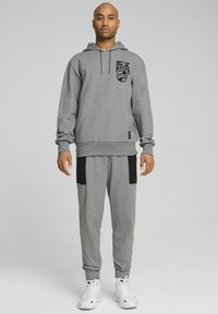 Sweat à capuche gris avec logo graphique noir, associé à un pantalon de jogging gris avec des accents noirs. Tissu lisse, poignets et ceinture côtelés. Baskets blanches.