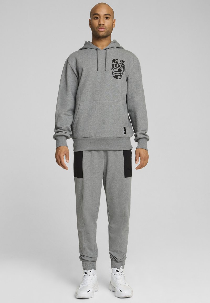 Sweat à capuche gris avec logo graphique noir, associé à un pantalon de jogging gris avec des accents noirs. Tissu lisse, poignets et ceinture côtelés. Baskets blanches.